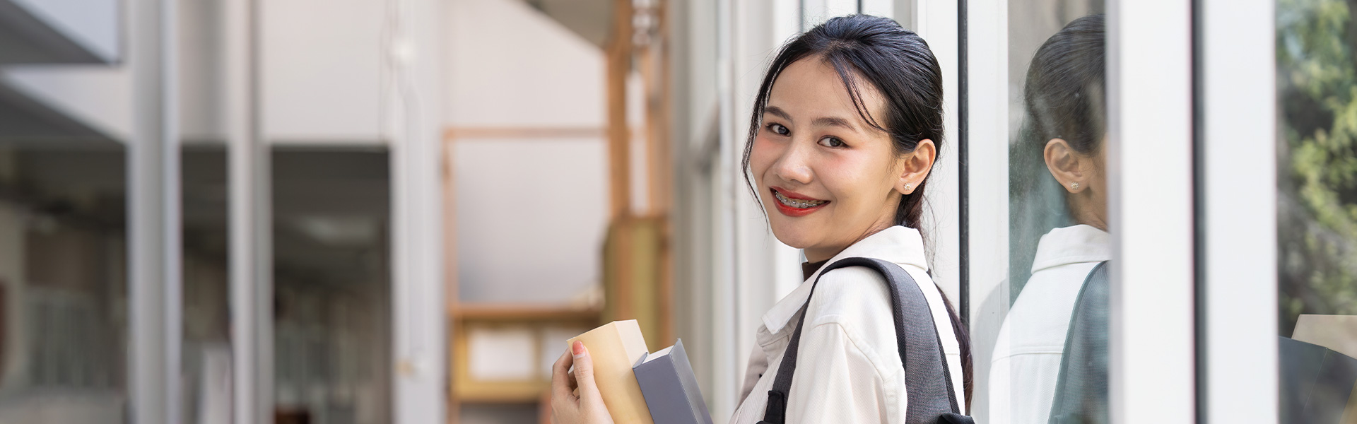 Asian female student holding books leaning against glass