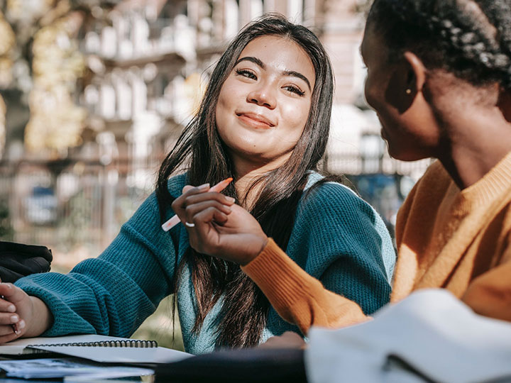 Two woman sitting next to each other at an outdoor seating area smiling at each other with notebooks open.