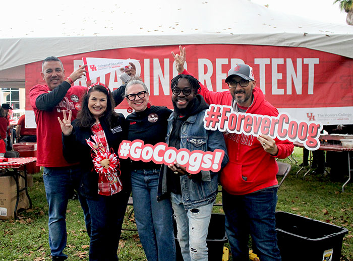 Four people at a tailgate event flashing Coog signs and holding signs that say Go Coogs and #ForeverCoog