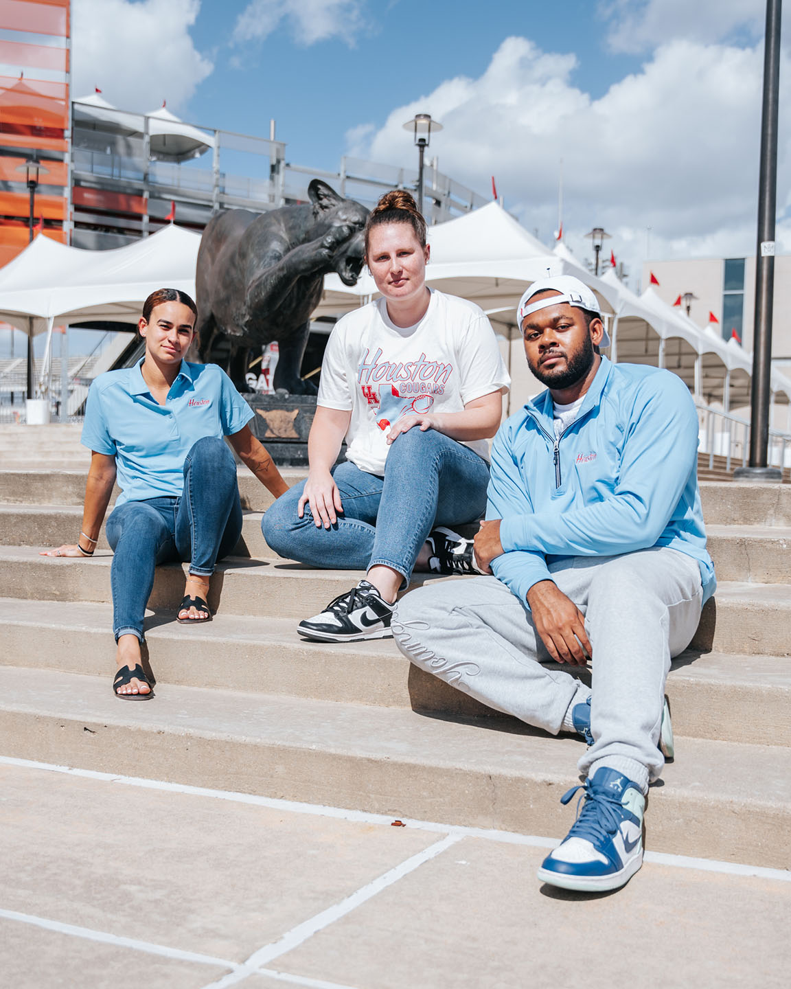 Three students sit together on the steps outside TDECU Stadium while wearing Houston Blue merchandise.