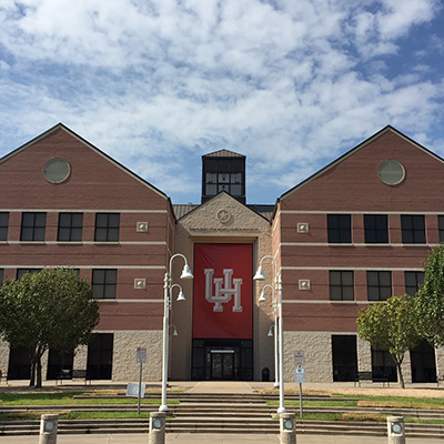 Photograph of a light brown brick building with a large red UH banner above the front entrance.