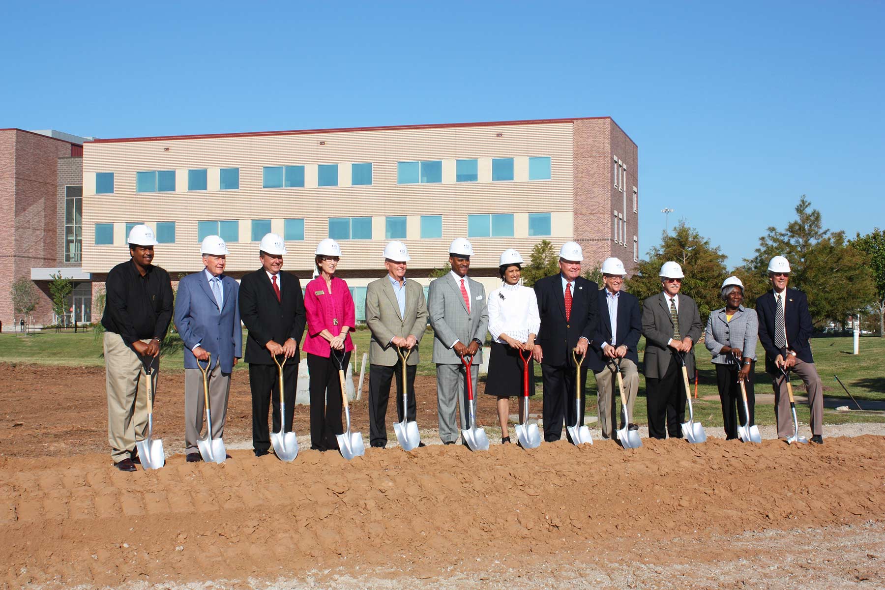 A ground of men and women in hardhats stand in a line holding shovels on a mound of dirt.