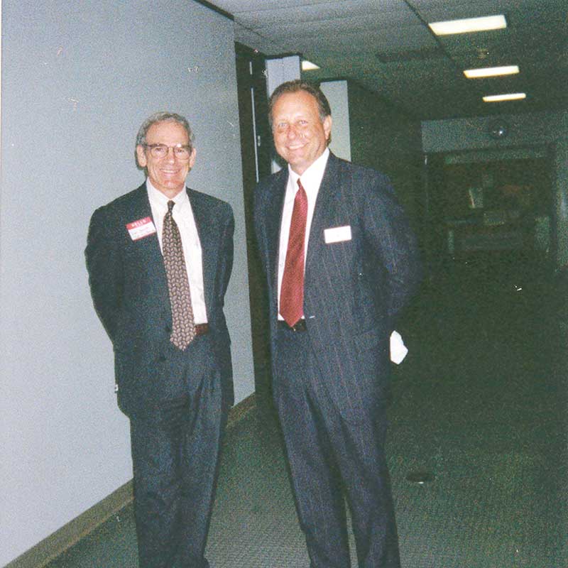 Photograph of two men in suits standing in a hallway.